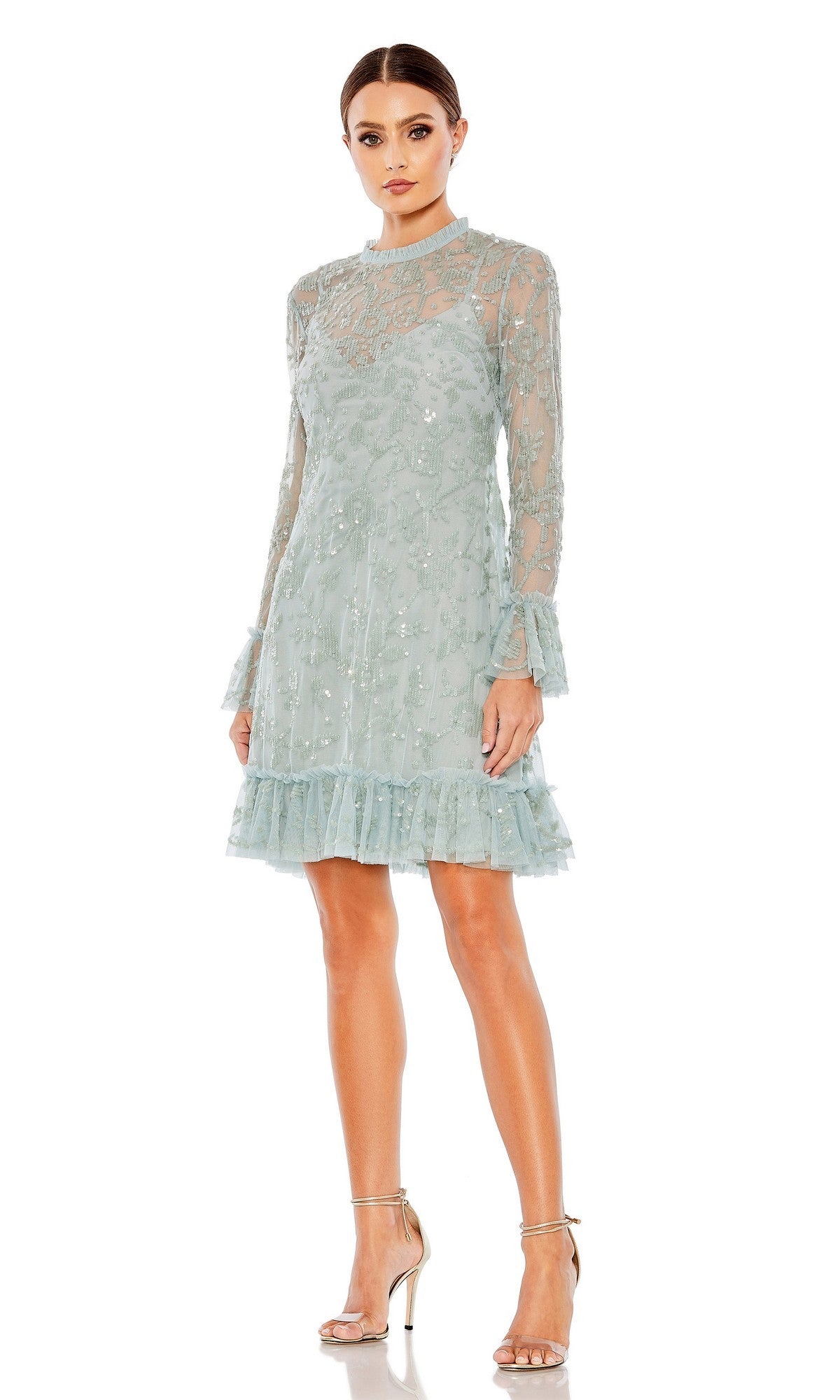 A woman wearing the Mac Duggal 9139 short party dress in seafoam—a mini cocktail dress featuring long sleeves, a high neckline, ruffled hem, and delicate sequin detailing—paired with silver high-heeled sandals, standing against a white background.